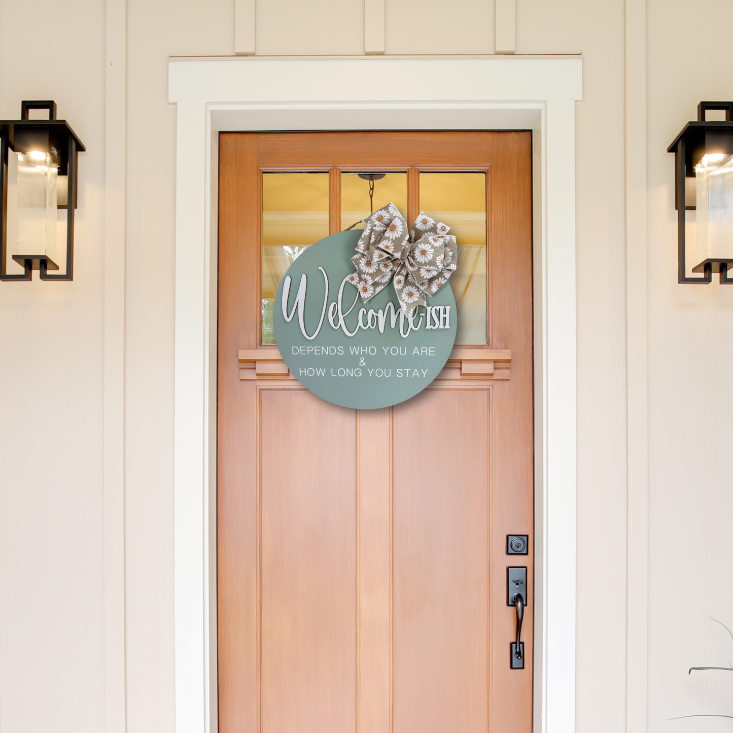 A round sage green welcome sign hanging on a wooden front door, decorated with a burlap and white daisy patterned bow. The sign reads 'Welcome depends who you are & how long you stay.