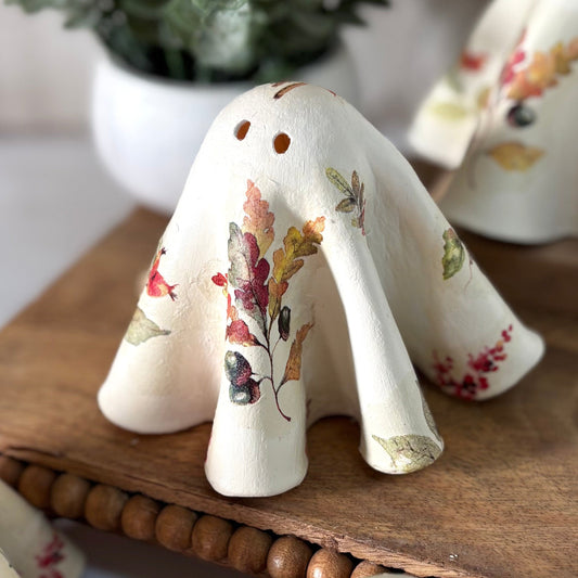 Set of ceramic ghosts with fall leaf patterns on a wooden stand, with a plant in the background.