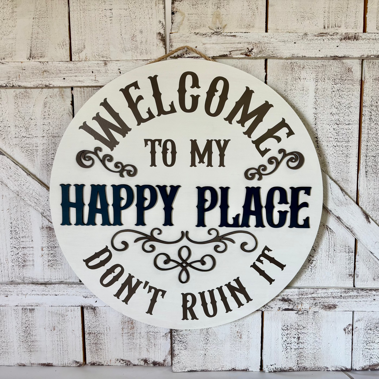 Round white wooden sign with black and navy lettering that reads 'WELCOME TO MY HAPPY PLACE DON'T RUIN IT,' hanging against a distressed white wood plank wall.
