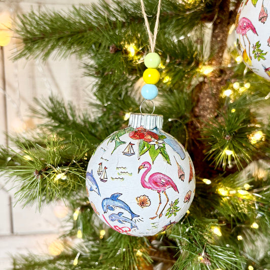 A close-up of a Christmas tree decorated with green pine branches, warm white string lights, and round Florida-themed ornaments. One ornament in the foreground shows colorful illustrations of flamingos, a blue dolphin, pelicans, and other sunshine state items on a light background.