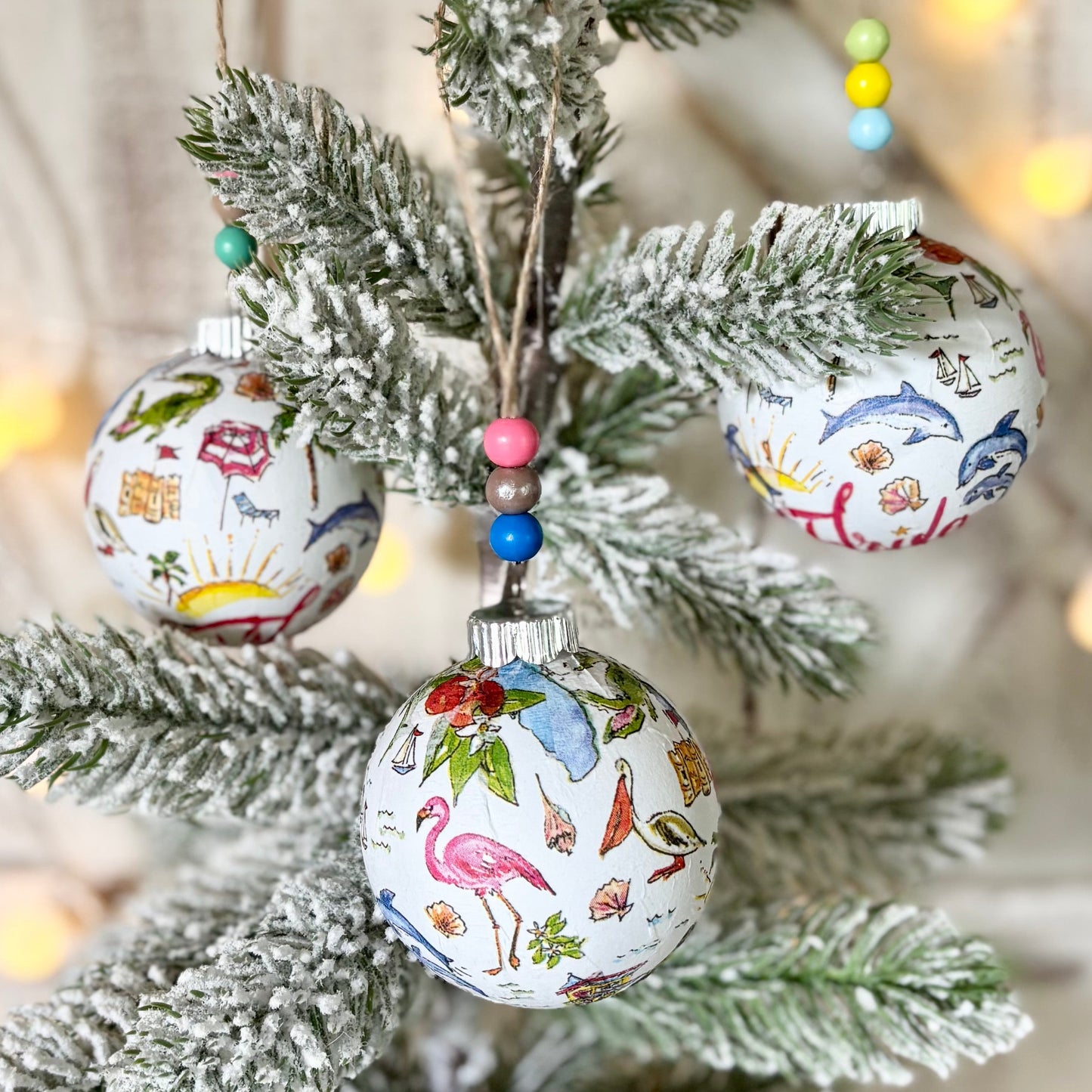 A small frosted artificial Christmas tree decorated with three patterned ornaments, set against a rustic white wood background with warm string lights.