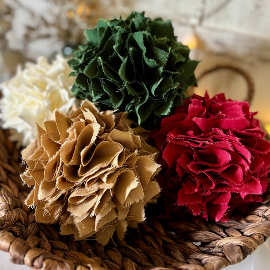 Four fabric pom-pom rag balls in green, red, tan, and cream, arranged in a round woven brown basket with warm string lights in the background.