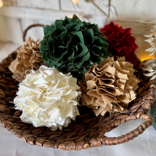 A set of decorative fabric rag ball bowl fillers in cream, green, red, and tan, arranged in a round woven basket on a white fabric surface with warm string lights in the background.