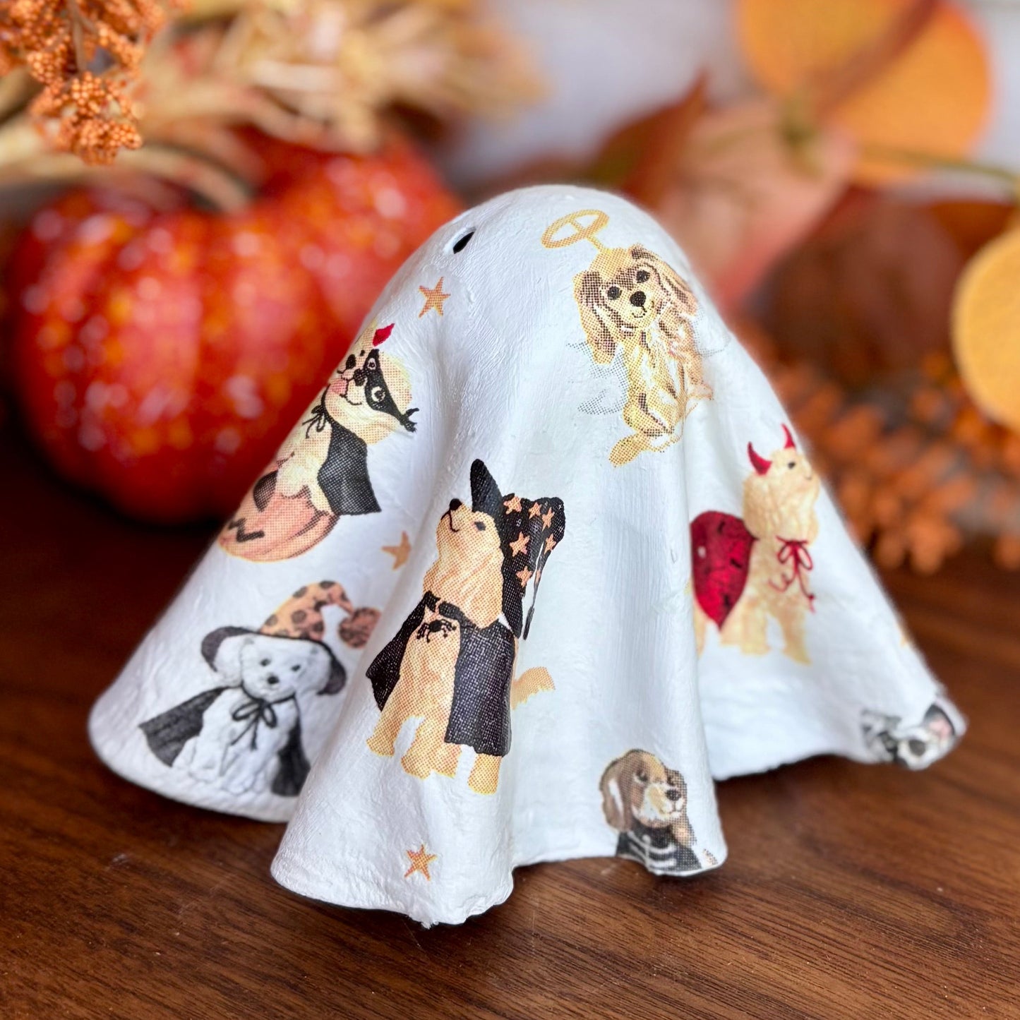 A white fabric ghost-shaped item printed with various cartoon dogs, displayed on a wooden surface next to autumn-themed decorative props including a small pumpkin and dried orange foliage.