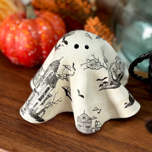 A decorative ceramic ghost figurine with a black and white toile pattern sits on a wooden serving board, styled with autumn fall decor including orange foliage, a small pumpkin, and a blue glass vase.