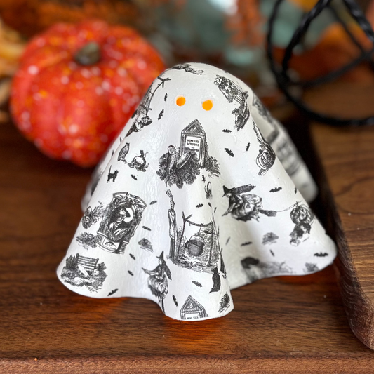 A small ghost-shaped ceramic figurine with a black and white witch and broom pattern sits on a wooden board, styled with autumn decorations in the background.