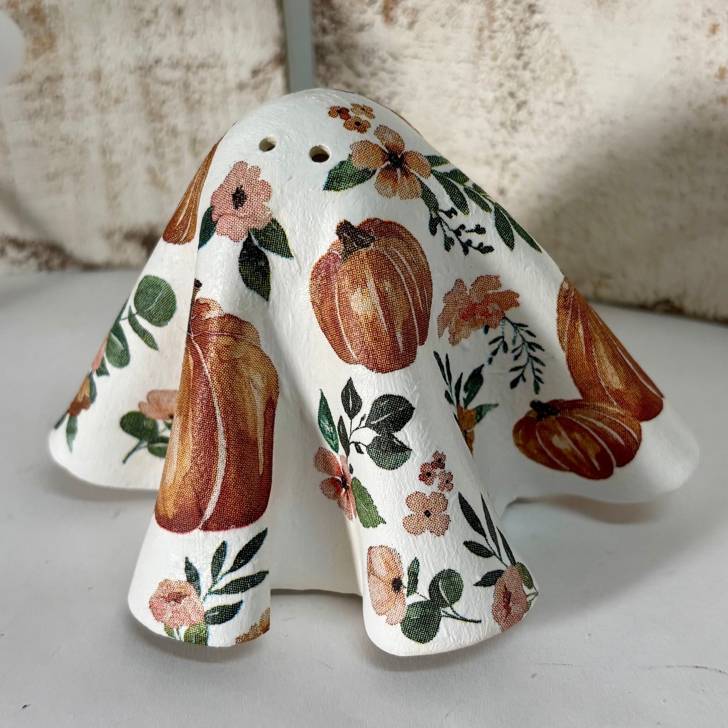 A white ceramic ghost, decorated with a rustic autumn pattern of orange pumpkins, green leaves, and pink flowers, displayed on a white surface against a distressed wood panel background with string lights.