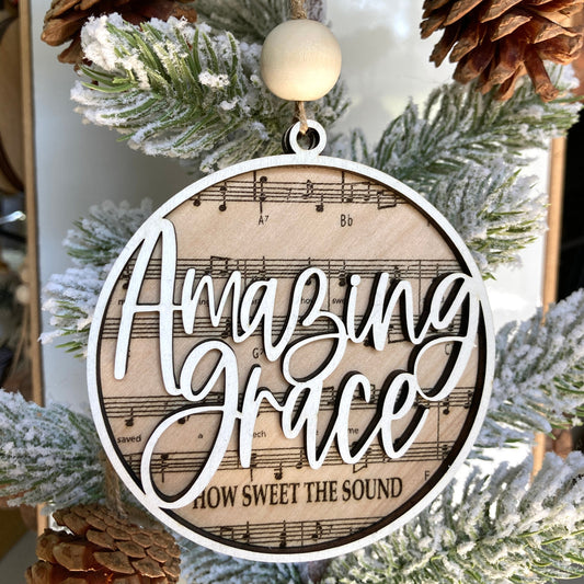 Large round ornament with Amazing Grace music notes laser engraved on natural wood. The words Amazing Grace is framed & cutout, painted white and applied on top of the wooden sheet music. Topped off with a large wood bead and jute cord