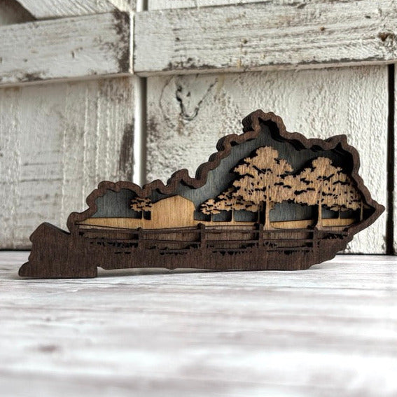Wooden cutout of a landscape with trees and a cabin on a wooden surface. Kentucky farm scene
