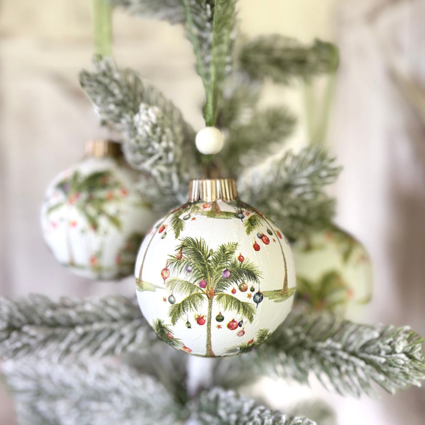 Round ornament repeating palm trees with red, blue and white ornaments hanging off of them on a white background, gold topper, green organza ribbon and white wood bead.
