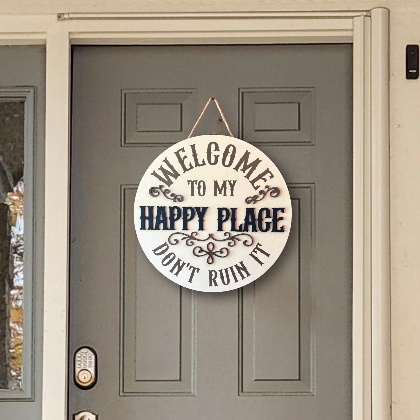 A round white wooden hanging sign with black text reading 'WELCOME TO MY HAPPY PLACE DON'T RUIN IT' displayed on a gray front door.