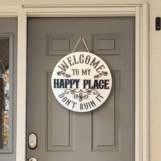 A round white wooden hanging sign with black text reading 'WELCOME TO MY HAPPY PLACE DON'T RUIN IT' displayed on a gray front door.