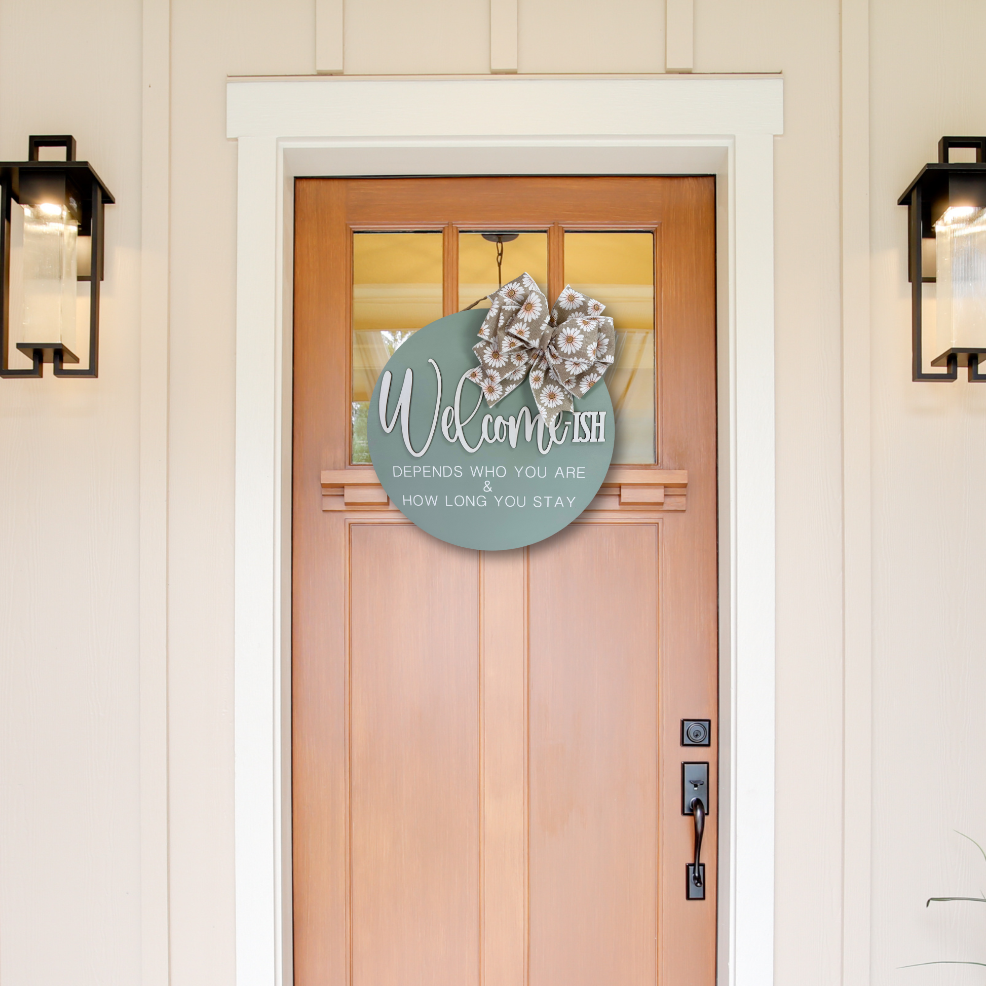A round sage green welcome sign hanging on a wooden front door, decorated with a burlap and white  daisy patterned bow. The sign reads 'Welcome depends who you are & how long you stay.