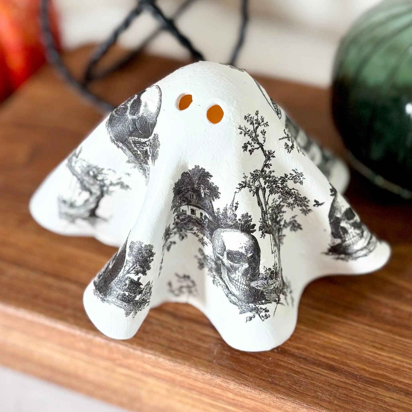 A ceramic ghost figurine with a black and white toile de Jouy pattern and tealight lit eyes, displayed on a wooden cutting board with seasonal props.