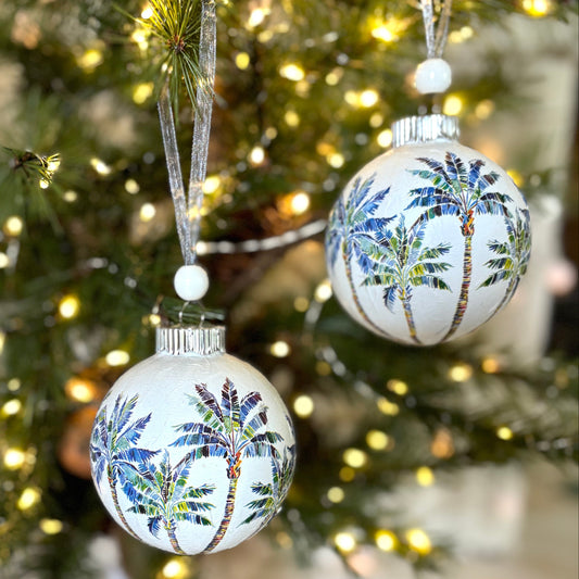 Decorative Christmas ornaments with palm tree patterns hanging in front of a lit Christmas tree.