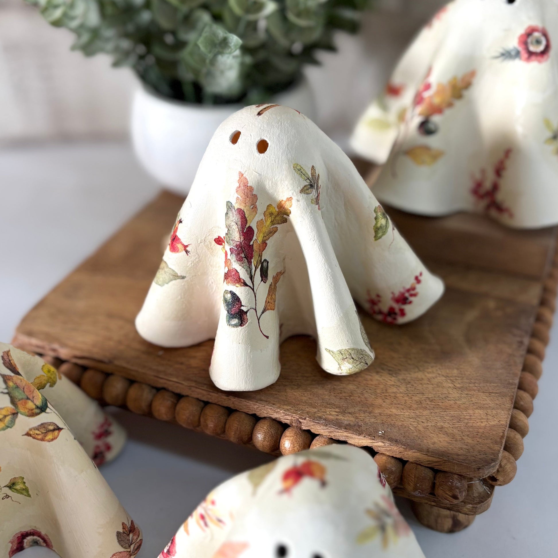 Set of ceramic ghosts with fall leaf patterns on a wooden stand, with a plant in the background.
