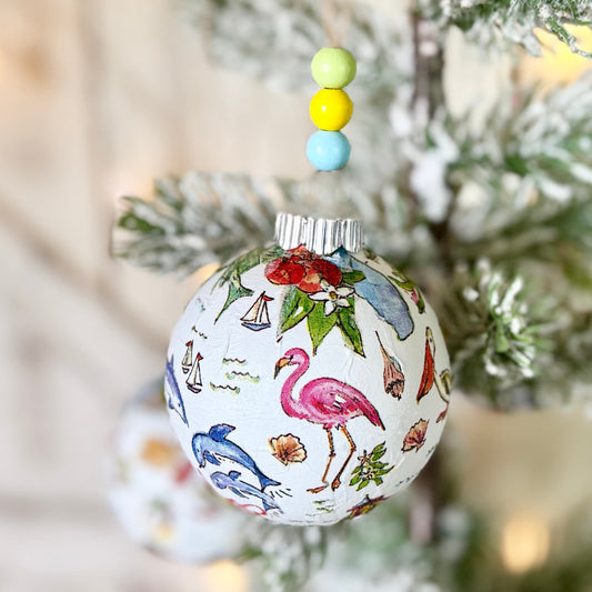 A decorative Christmas ornament featuring a colorful flamingo and bird pattern on a white background, hanging on a frosted pine tree branch with small pastel beads above it.