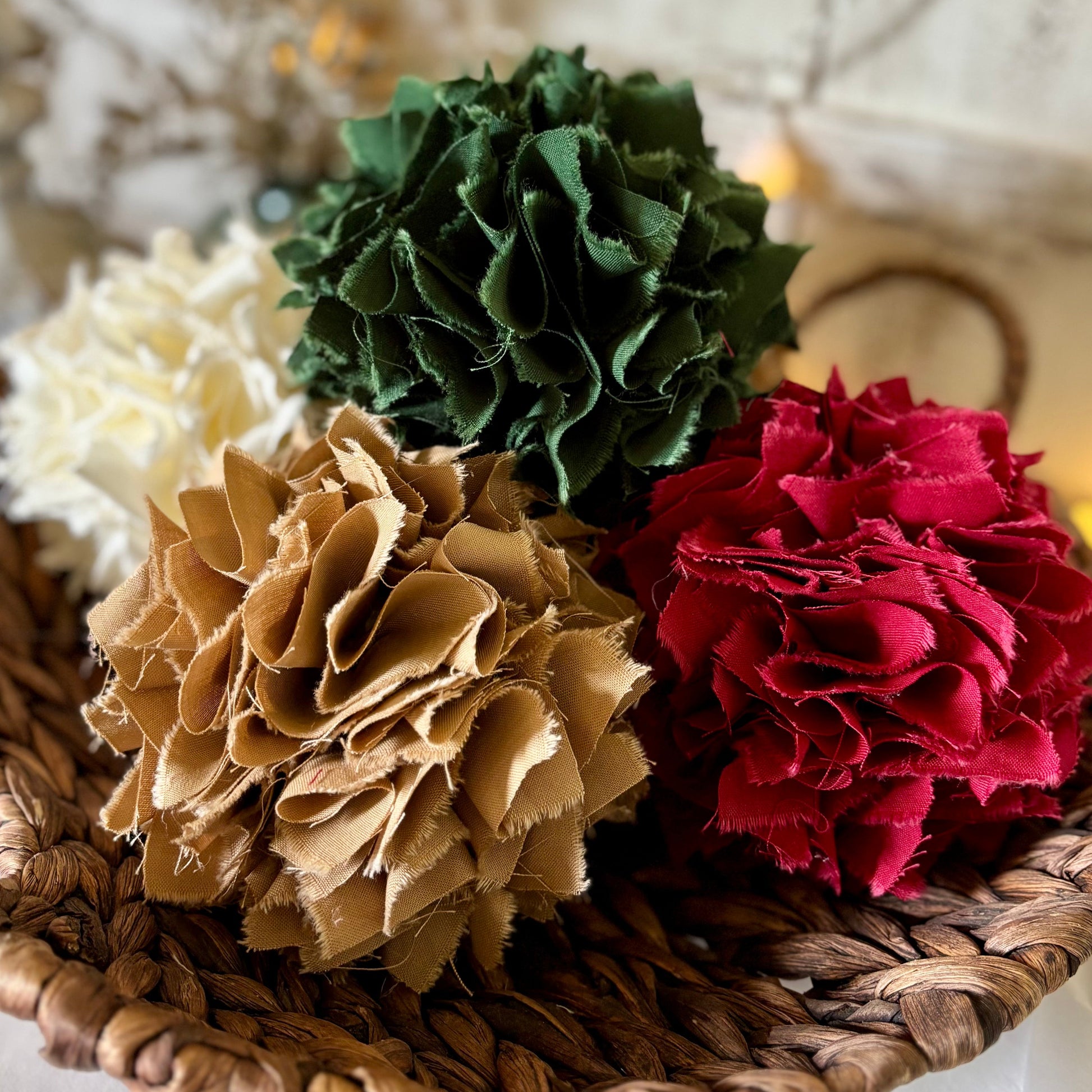 Four fabric pom-pom rag balls in green, red, tan, and cream, arranged in a round woven brown basket with warm string lights in the background.