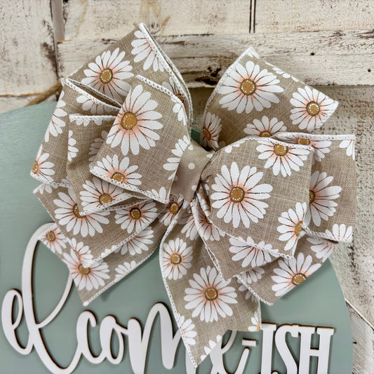 A large fabric bow with a white daisy floral print on a beige linen-like background, displayed on a rustic white wooden sign that reads "Welcome-ish" against a distressed whitewashed wood backdrop.