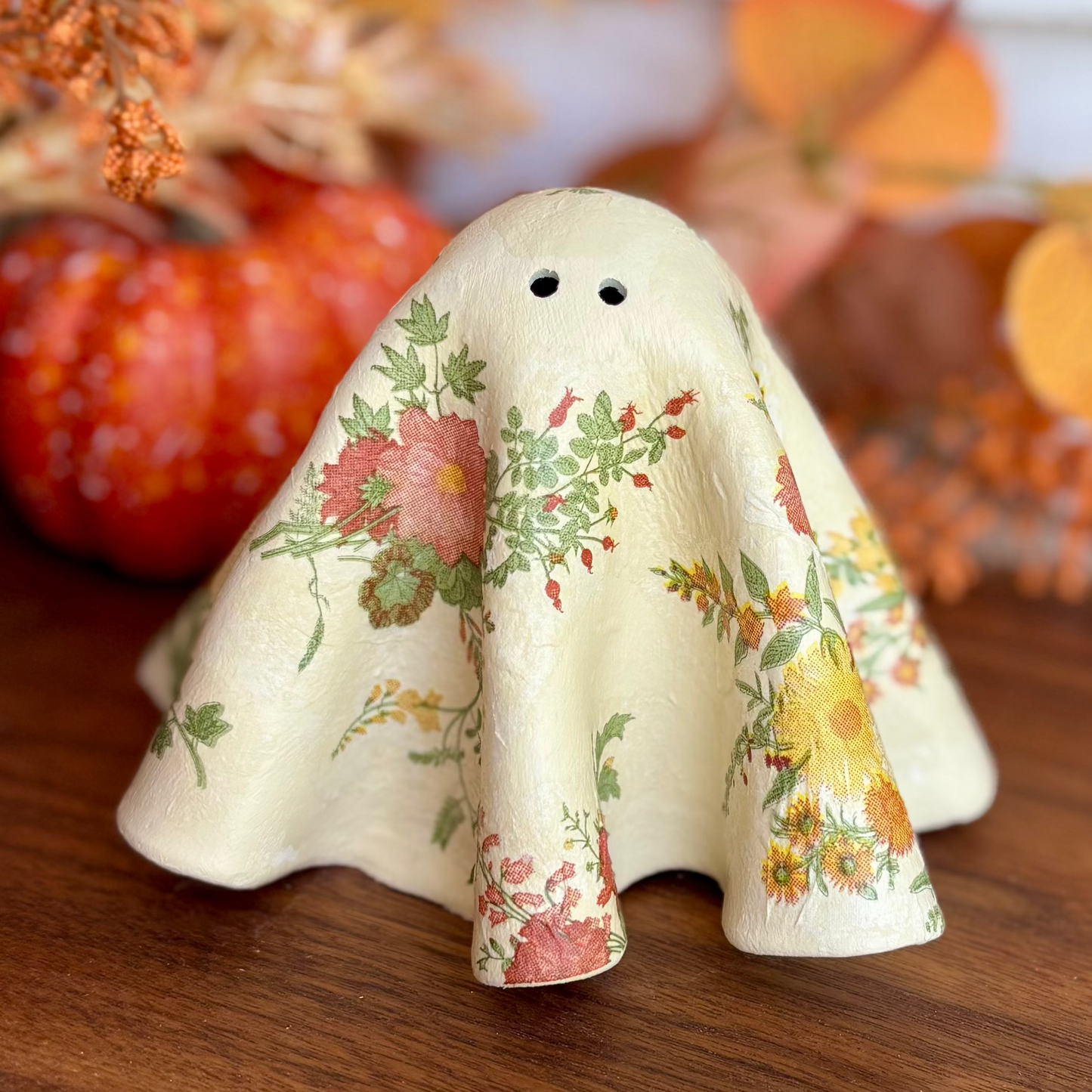 A small decorative ghost figurine with a cream-colored fabric-like body and a multicolor floral print, styled on a wooden surface with autumn leaves and a small pumpkin in the background.