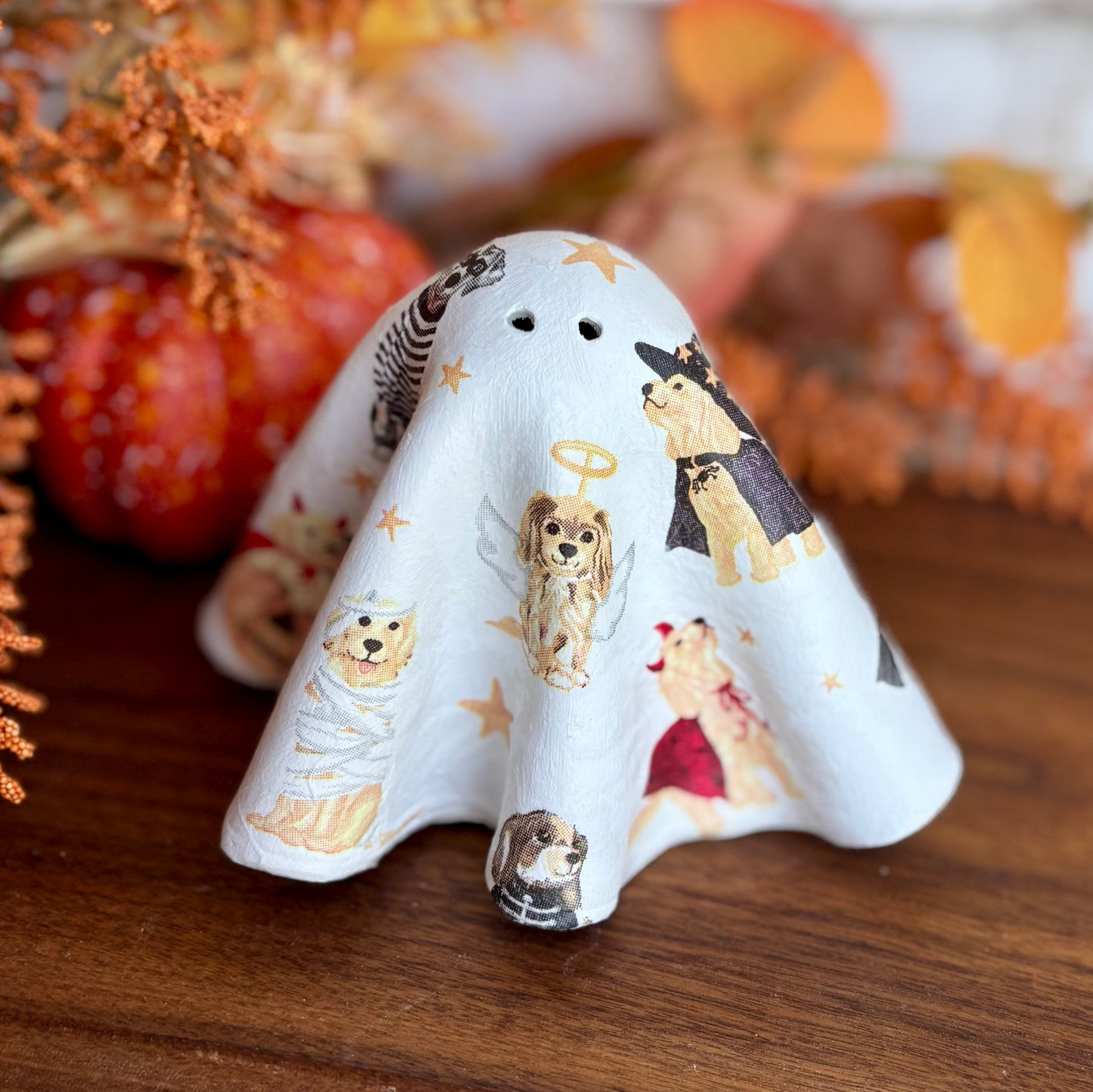 A small white ghost-shaped ceramic figurine decorated with cute cartoon dogs in different outfits, displayed on a wooden surface with autumn-themed decorative props in the background.