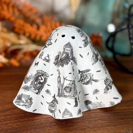 A ghost-shaped ceramic decoupaged with witches and graveyard pattern in a black and white Halloween print, displayed on a wooden table with autumn decorations in the background.