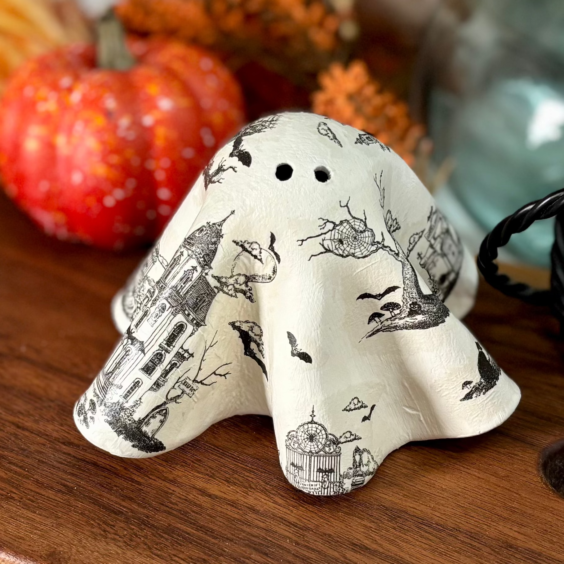 A decorative ceramic ghost figurine with a black and white toile pattern sits on a wooden serving board, styled with autumn fall decor including orange foliage, a small pumpkin, and a blue glass vase.