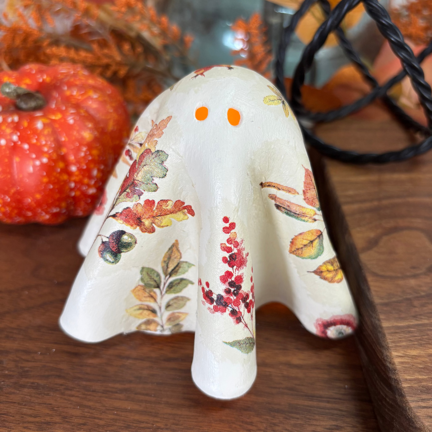 A decorative ghost figurine with an off-white base and a decoupaged autumn leaf and berry pattern, displayed on a wooden surface with seasonal props.