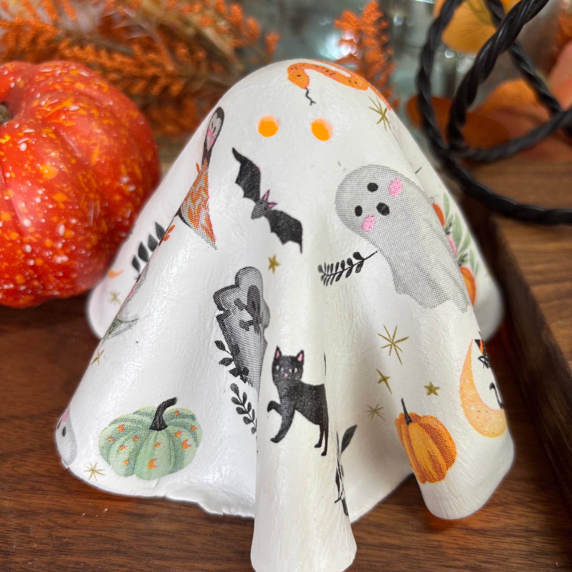 A white Halloween-themed ghost-shaped candle holder with printed illustrations of black cats, orange pumpkins, a gray ghost, bats, and stars, displayed on a wooden surface with autumn decorations.