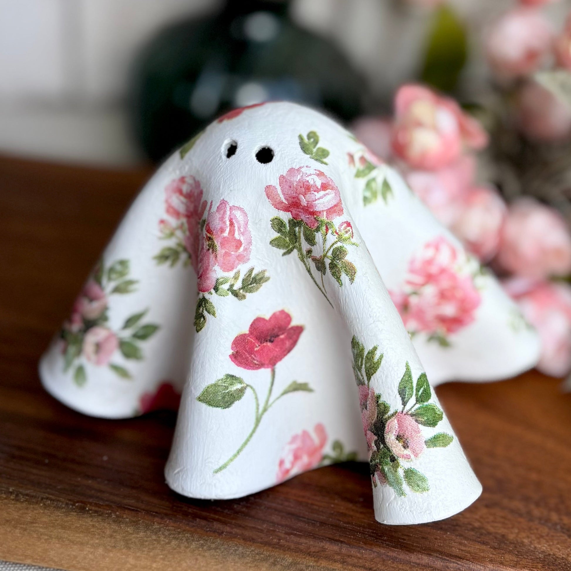 A small white ceramic ghost figurine with a pink and green floral print, displayed on a wooden board with dried flowers in the background.