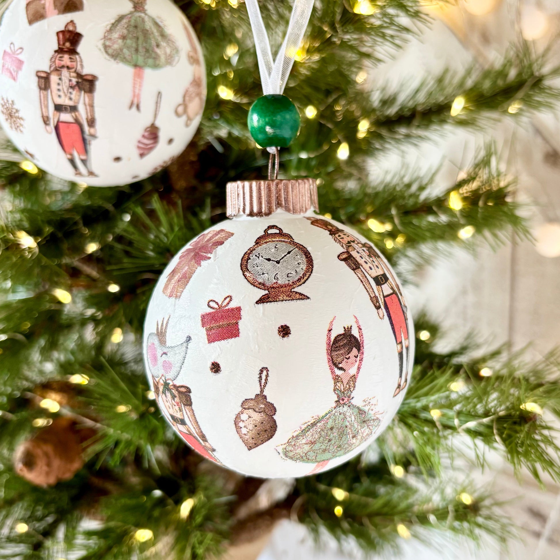Two round Christmas ornaments with vintage illustrations of ballerinas and other holiday motifs, hanging on a decorated Christmas tree with warm string lights.