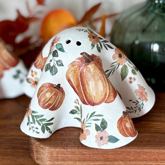 Two white ceramic ghosts figurines decorated with orange pumpkins and green leaves, displayed on a wooden cutting board with a green glass vase and dried leaves in the background.