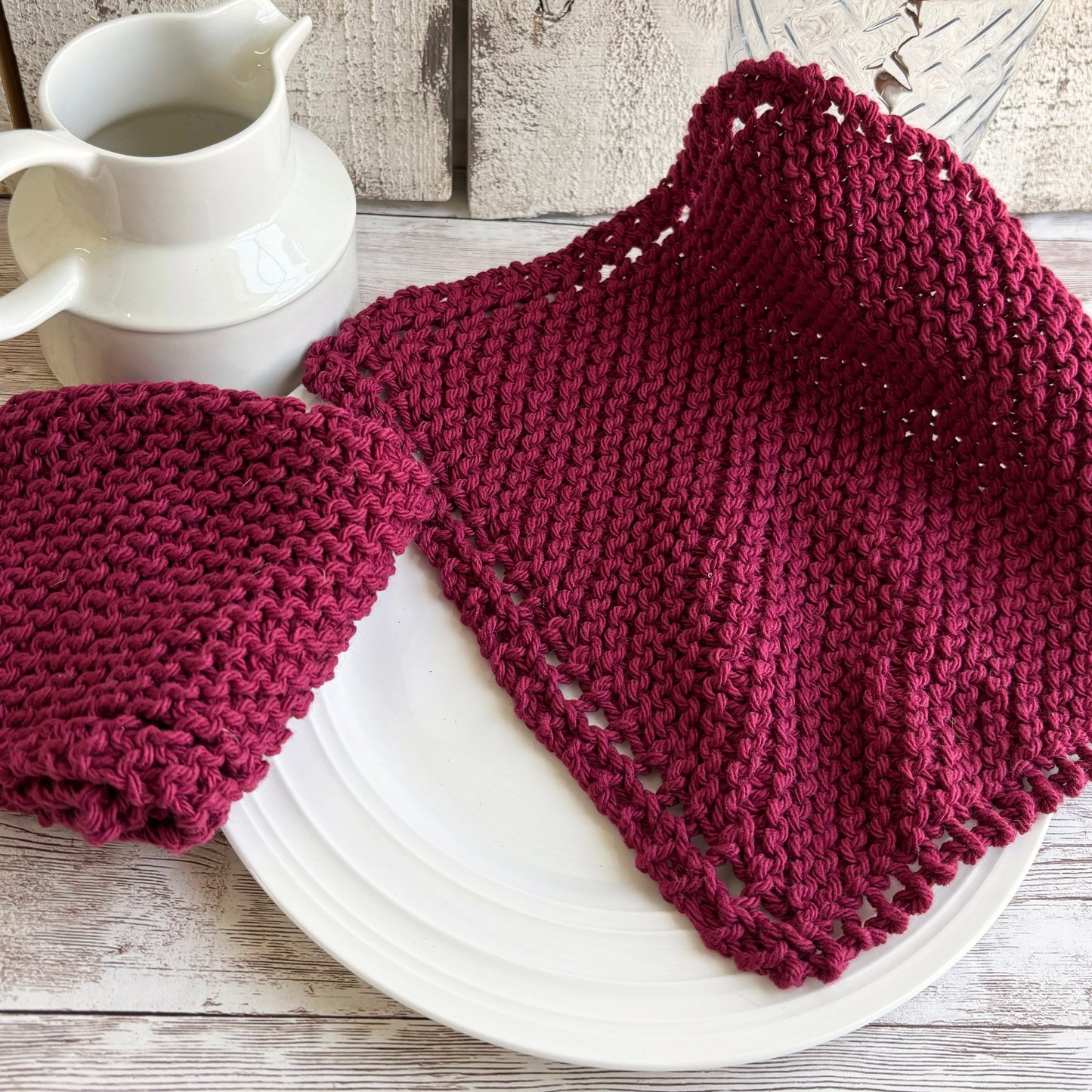 Knitted maroon dishcloths on a white plate with a white pitcher and glass against a wooden background
