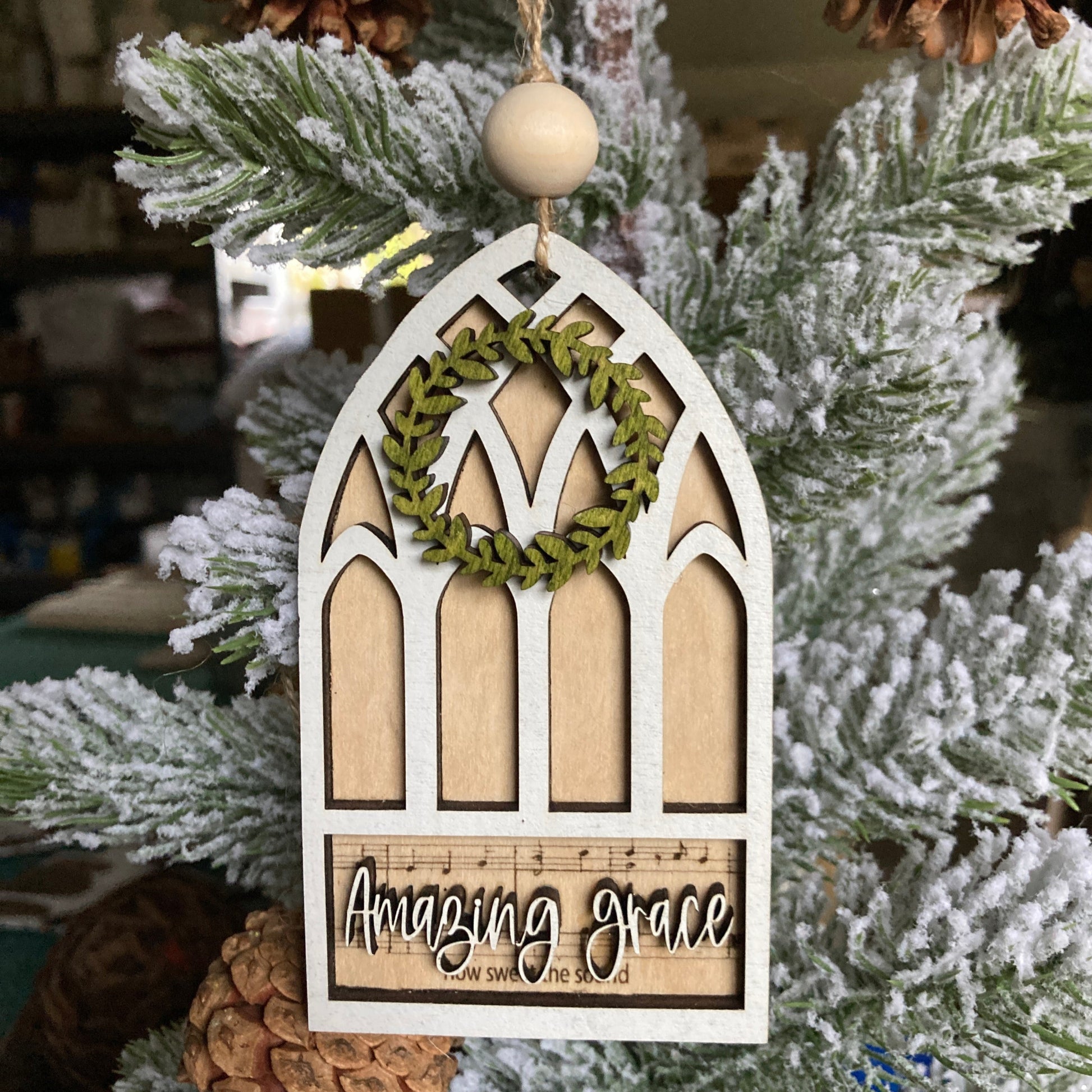 Stained glass window shaped ornament that has Amazing Grace sheet music laser engraved on natural wood for the background. The top layer is the window shape with a green wooden wreath at the top. A wood bead is with the jute string hanger