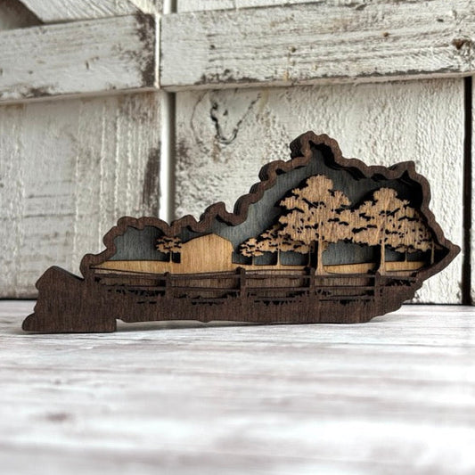 Wooden cutout of a landscape with trees and a cabin on a wooden surface. Kentucky farm scene