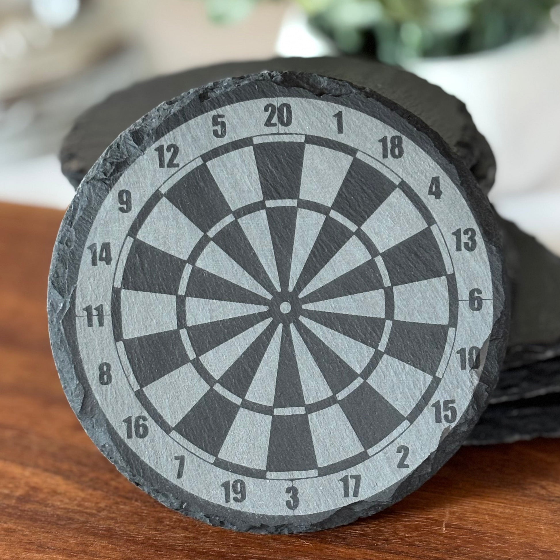 Black slate coaster with a Dartboard design on a wooden surface with a blurred background
