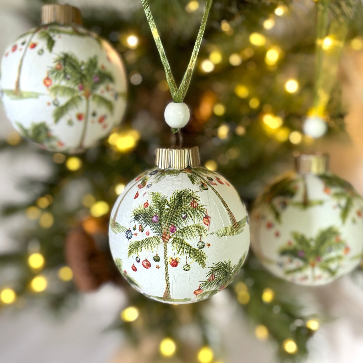 Round ornament repeating palm trees with red, blue and white ornaments hanging off of them on a white background, gold topper, green organza ribbon and white wood bead.