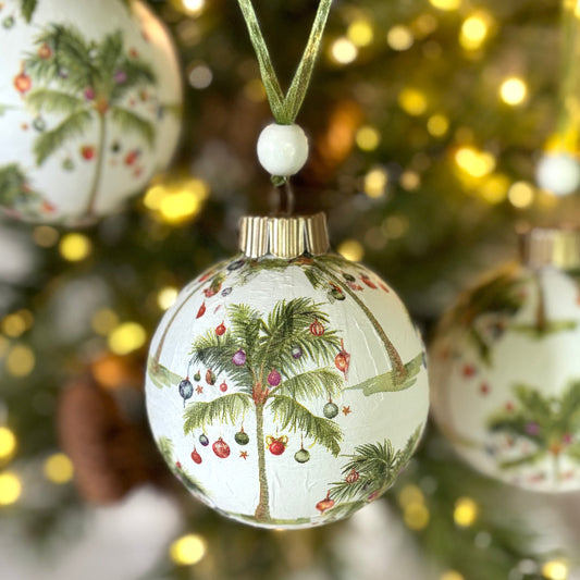 Round ornament repeating palm trees with red, blue and white ornaments hanging off of them on a white background, gold topper, green organza ribbon and white wood bead.