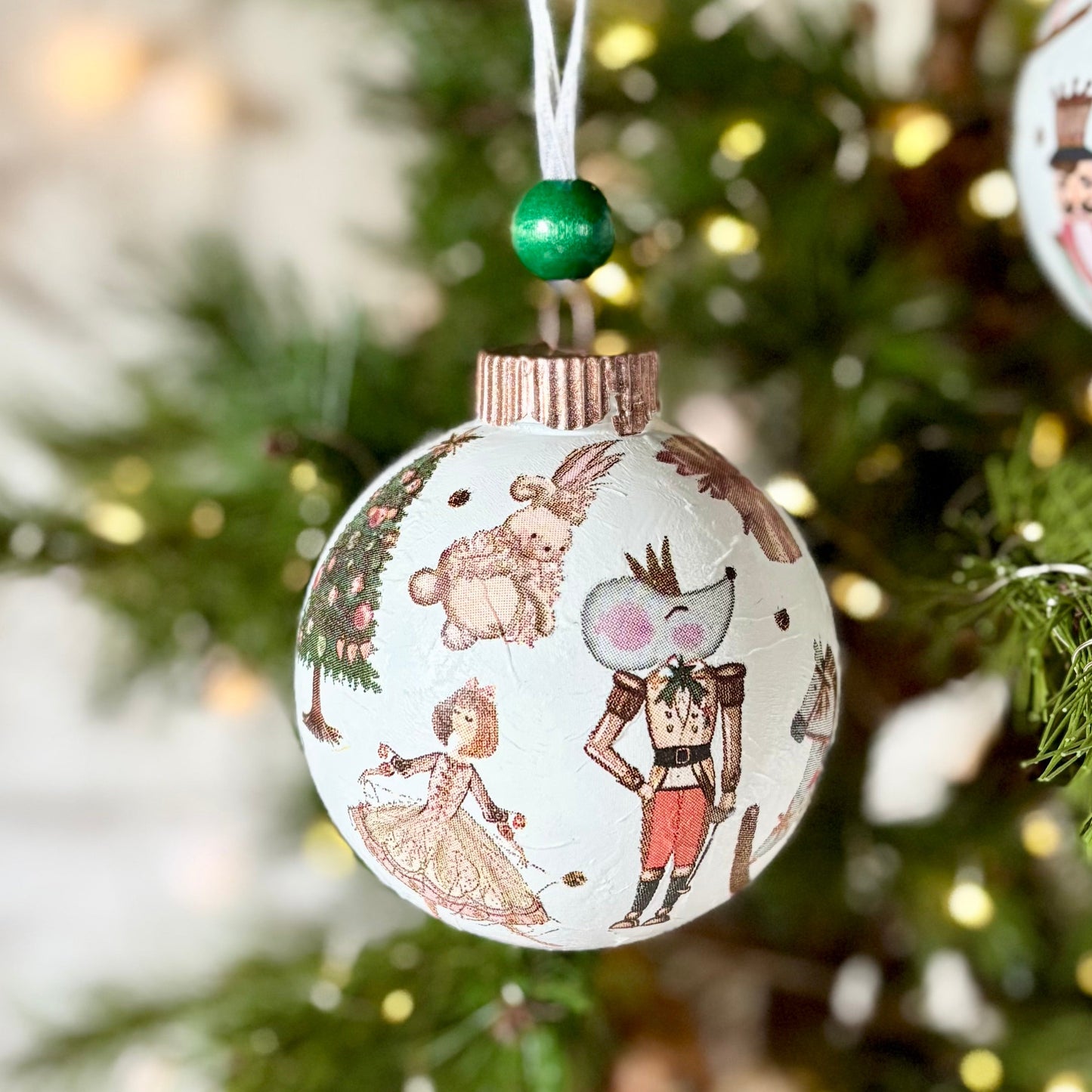 Two round Christmas ornaments with vintage-style illustrated patterns hang on a decorated evergreen tree with warm string lights.