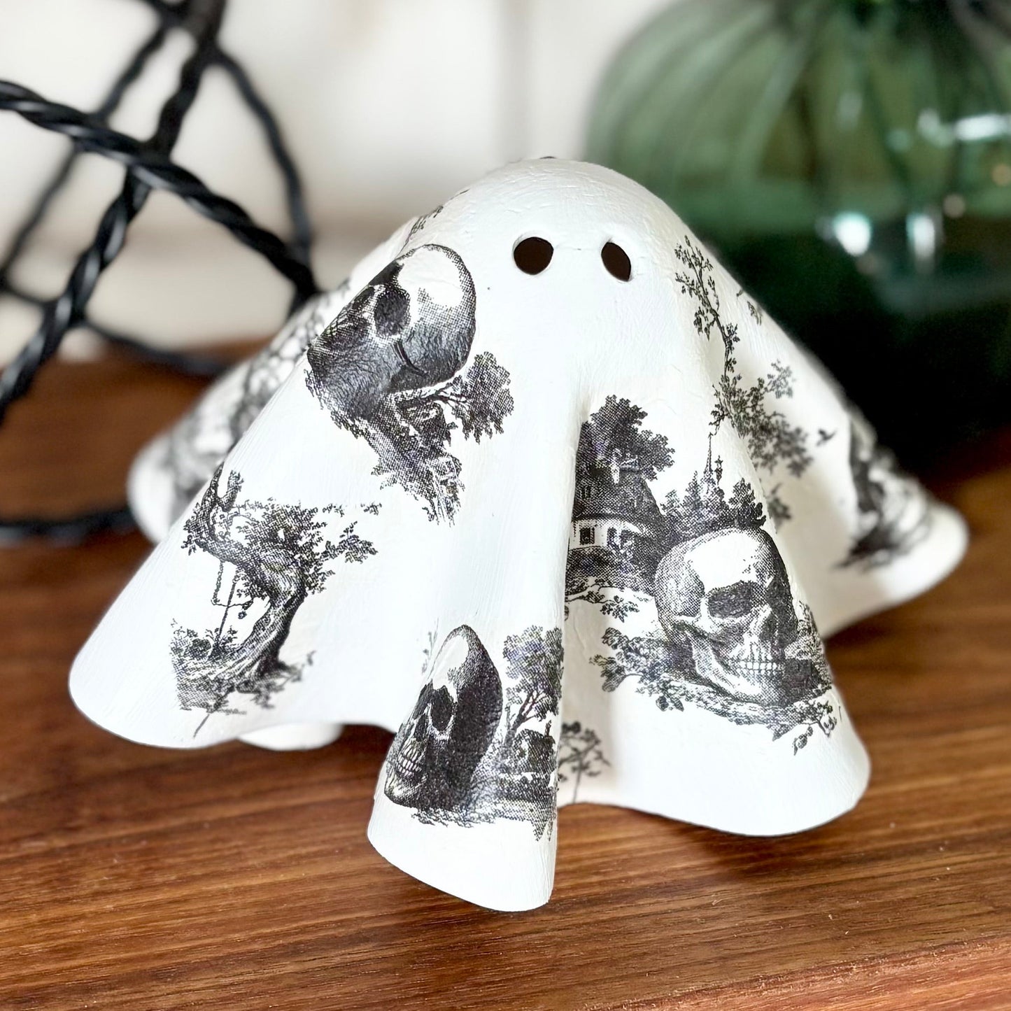 A white ceramic ghost-shaped clay figurine with a black gothic illustration skull pattern, displayed on a wooden cutting board with seasonal props.